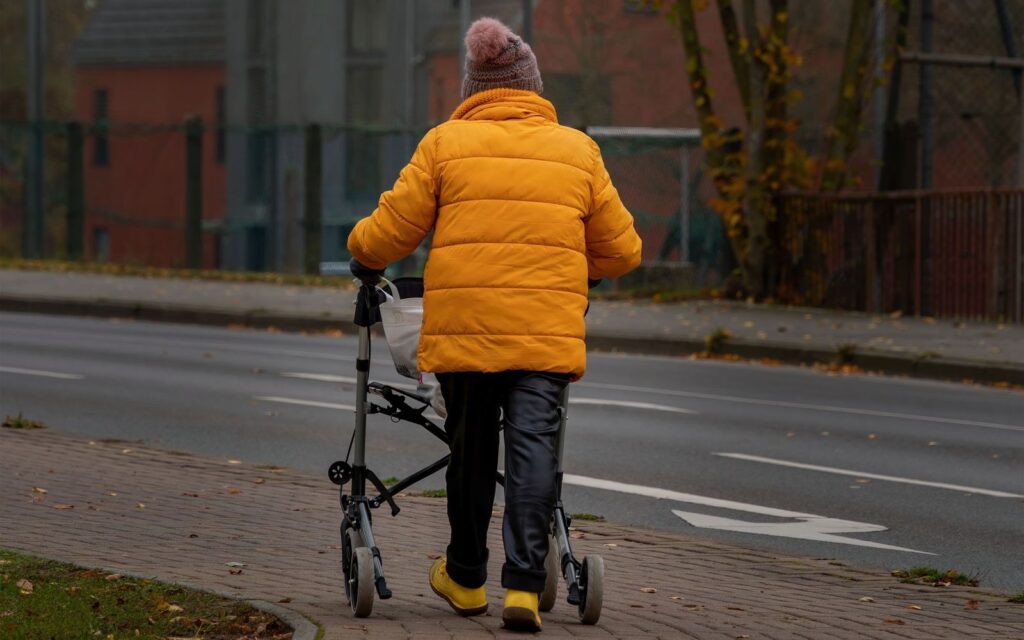 Older adult walking outdoors with a rollator on a tree-lined sidewalk